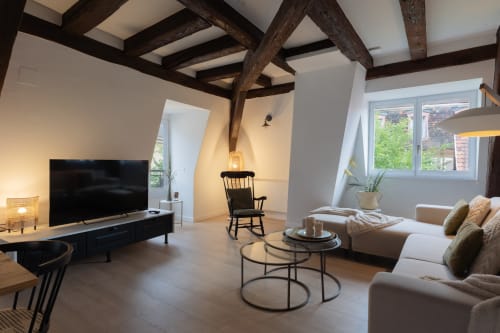Modern loft-style living room at Le JO·LIE Duplex in Colmar with exposed beams, beige sofa, TV, and rocking chair