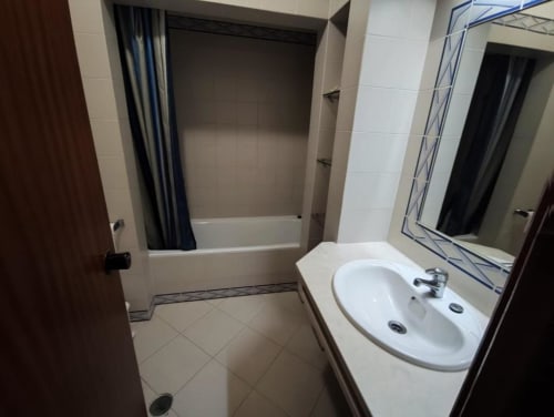 Banheiro moderno no apartamento Albufeira Inn com pia branca, espelho e banheira com cortina de chuveiro azul