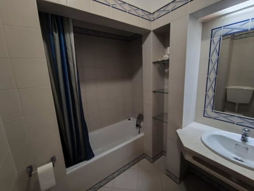 Banheiro moderno do apartamento Albufeira Inn com banheira, cortina de chuveiro, pia, espelho e paredes com azulejos brancos