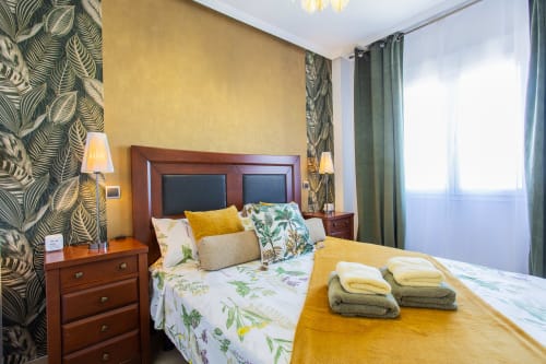 Luminoso dormitorio en Haus Holiday Homes con cama de madera, ropa de cama floral, paredes amarillas, papel tapiz tropical y vista a la piscina en San