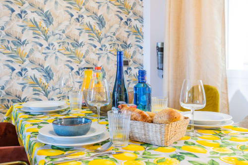 Mesa de comedor preparada con mantel de estampado de limones amarillos, copas de vino, cesta de pan y botellas en apartamento Haus Holiday Homes