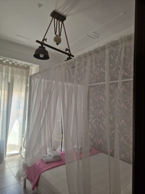 Cama com dossel e cortinas brancas translúcidas no quarto de hóspedes da Zuni Guest House Lisboa, com roupa de cama rosa e papel de parede estampado