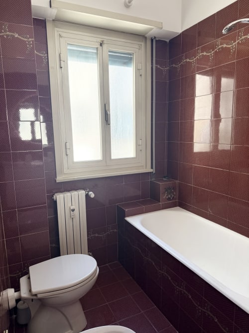 Modern bathroom at Claudia Apartments Rome with burgundy tiled walls, white bathtub, toilet, heated towel rail, and window