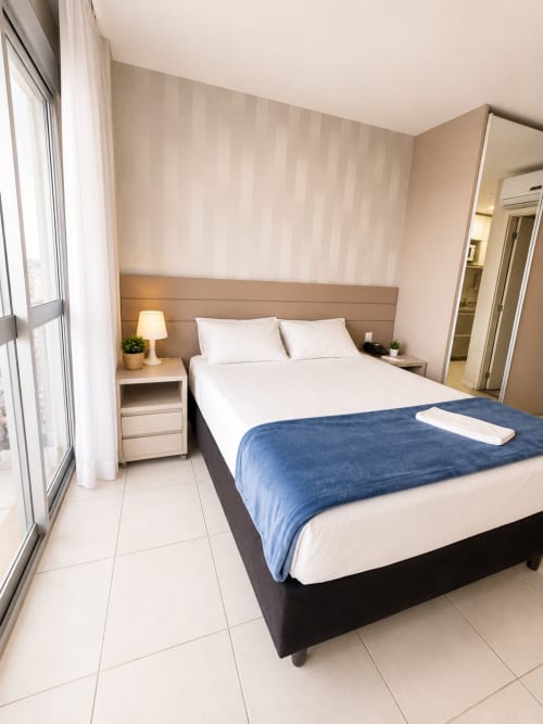 Quarto moderno em pousada de Brasília com colcha azul, linhos brancos, cabeceira em madeira, abajur e vista da cidade pela janela
