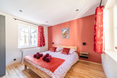 Dormitorio moderno en Les Ambrades de Kaysersberg con paredes coral, cama de madera, acentos rojos y grandes ventanales