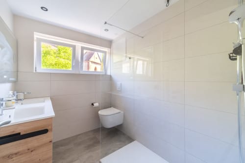 Baño moderno con azulejos blancos, inodoro flotante, mueble de madera y vistas por ventana en apartamento de Kaysersberg