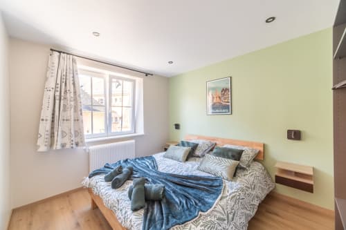 Dormitorio luminoso con paredes verde pálido, cama de madera, ropa de cama estampada y vistas por la ventana en el apartamento Les Ambrades, Kaysersberg.