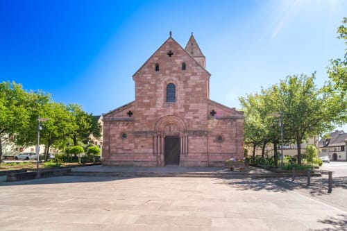 Iglesia de arenisca roja con dos torres en la soleada plaza de Kaysersberg Vignoble, rodeada de árboles verdes y edificios