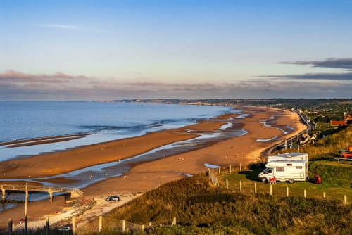 Camping-car à Skol en Normandie surplombant une plage de sable doré avec motifs de marée et falaises côtières