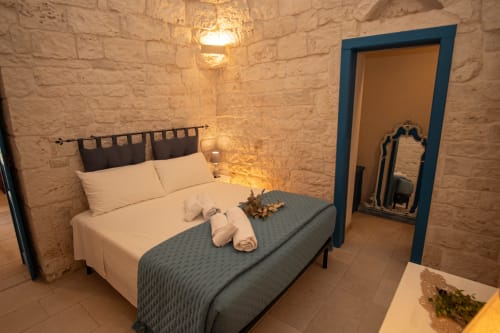 Accogliente camera da letto in trulli a Ostuni con muri in pietra, biancheria bianca, infissi blu scuro e illuminazione calda