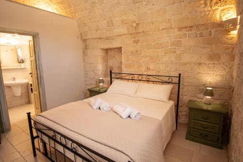 Camera Trulli Life a Bari con muri in pietra, letto in ferro battuto, biancheria color crema e comodini verdi