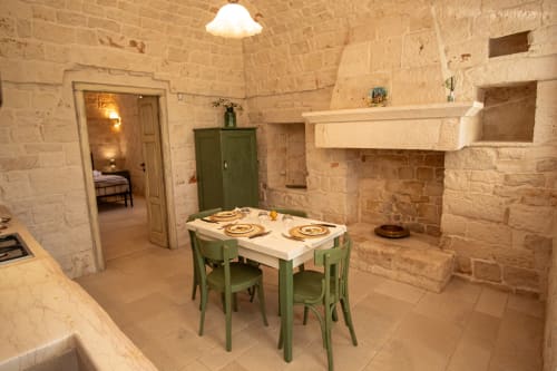 Area dining di Trulli Life con muri in pietra, tavolo e sedie verdi, camino e porta della camera da letto a Bari