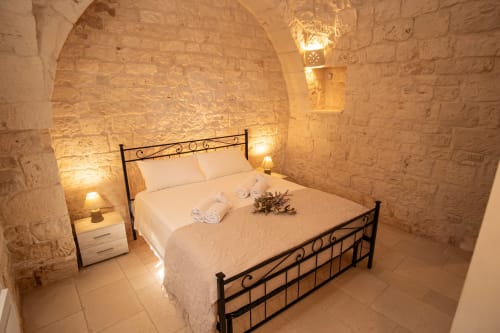 Camera da letto Trulli Life a Ostuni con muri in pietra, letto in ferro battuto, biancheria bianca e illuminazione ambientale