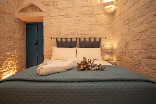Camera da letto Trulli Life a Ostuni con muri in pietra, porta in legno scuro e comodo letto con cuscini color crema