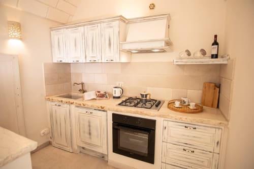 Cucina compatta con mobili bianchi, rivestimento in piastrelle beige, piano cottura a gas e scaffali aperti presso l'hotel Trulli Life di Ostuni