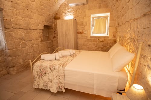 Camera da letto Trulli Life a Ostuni con muri in pietra, biancheria bianca, testiera in legno e illuminazione calda nella Terra Rossa Trullamie