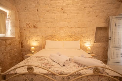 Camera da letto accogliente in Trulli Life Ostuni con muri in pietra, letto in ferro battuto, biancheria bianca e illuminazione calda