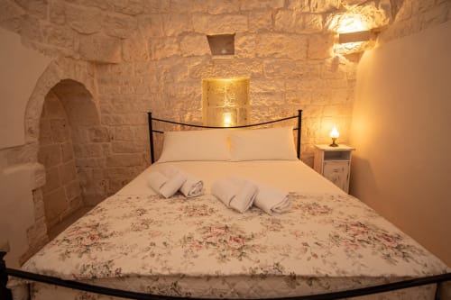 Accogliente camera in pietra ai Trulli Life di Ostuni con biancheria floreale, letto in ferro battuto nero e illuminazione ambiente calda
