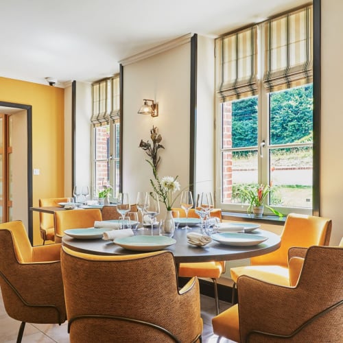 Modern dining room at Clos Céleste with round table, orange chairs, and large windows overlooking Etretat countryside.