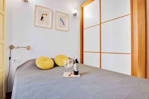 Modern bedroom at Ayma Apartamento Ollerías in Málaga with grey bed, yellow cushions, wine setup, and wooden window frames