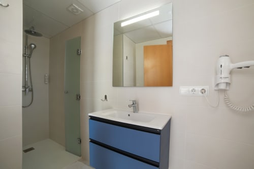 Cuarto de baño moderno en Apartamentos Playa Sol con vanidad azul, espejo, ducha y paredes de azulejos blancos en Es Canar