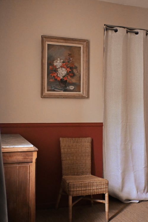 Coin élégant de chambre au Mas De La Digue avec tableaux floraux encadrés, fauteuil en osier et rideaux crème
