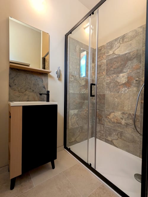 Salle de bains moderne du gîte Domaine Alga avec douche vitrée, murs en pierre, et vasque noire