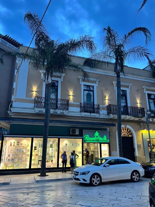 Palazzo sul Corso apartment storefront in Brindisi's historic city center at dusk with palm trees and white Mercedes parked