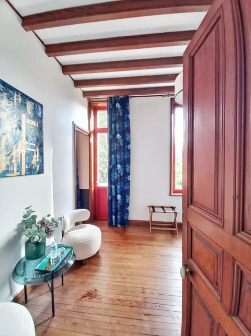 Elegant bedroom at Villa Eliane with exposed beams, hardwood floors, white chair, blue curtains, and red-framed windows