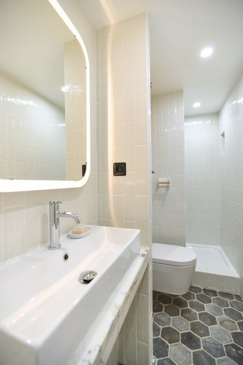Salle de bain moderne au Boutik Boheme Paris avec carrelage blanc brillant, miroir rétroéclairé, lavabo, toilettes et douche spacieuse