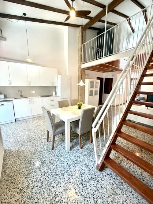 Moderna cocina-comedor tipo loft en Valencia con mueblería blanca, suelo de terrazo y escaleras de madera