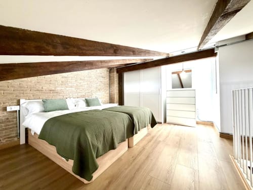 Moderno dormitorio loft en Valencia con vigas de madera vista, ropa de cama verde, paredes de ladrillo y suelos de madera