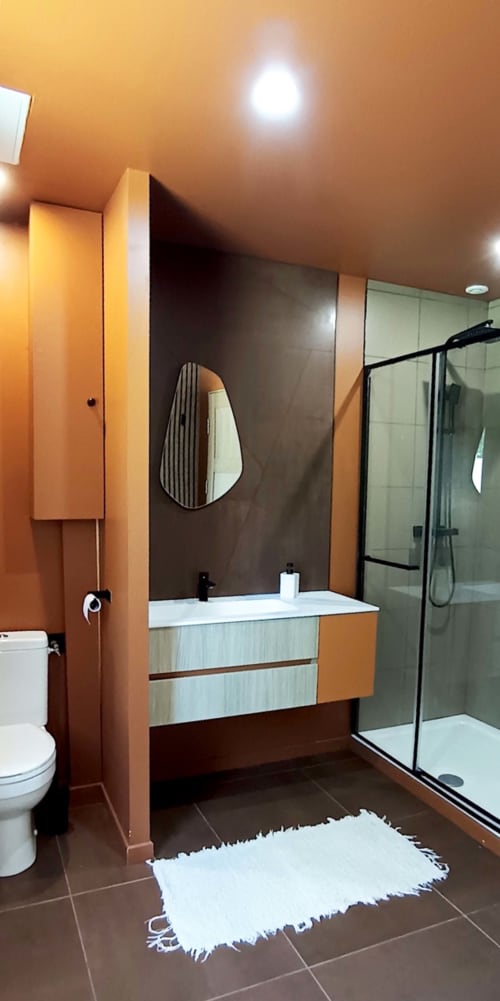 Salle de bain moderne du Domaine Alga avec murs accentués d'orange, vasque suspendue, miroir ovale et douche vitrée