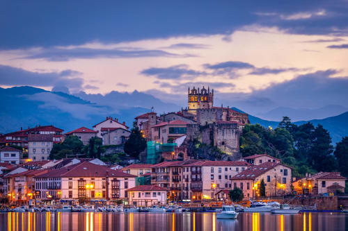 Twilight view of Comillas waterfront with illuminated historic buildings and castle tower reflected in calm water, Spain