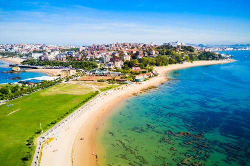 Aerial view of Comillas beach with golden sand, turquoise water, and coastal town nestled against green hills under blue sky