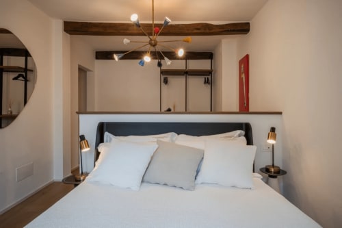 Camera moderna a Manu Maison di Parma con biancheria bianca, testiera scura, travi a vista e illuminazione artistica.
