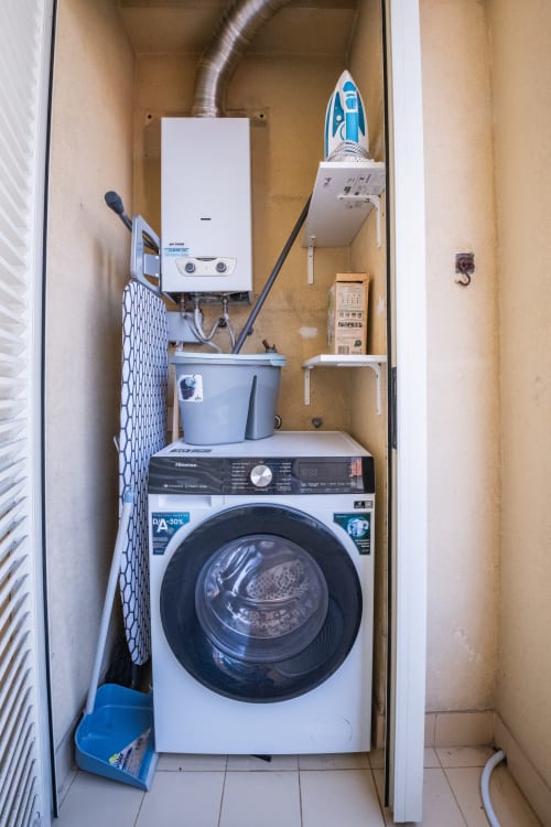 Kompakte Waschküche mit Waschmaschine, Warmwasserspeicher, Bügeleisen und Regalen im EcoHost Apartment