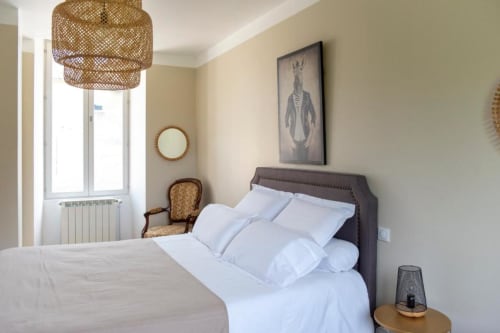 Chambre élégante à La Demeure du Comte avec literie blanche, tête de lit foncée, fenêtre sur le jardin et luminaire suspendu tissé