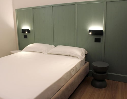 Camera da letto Maison Riviera a Reggio di Calabria con biancheria bianca, testata verde, applique moderne e pavimento in legno.