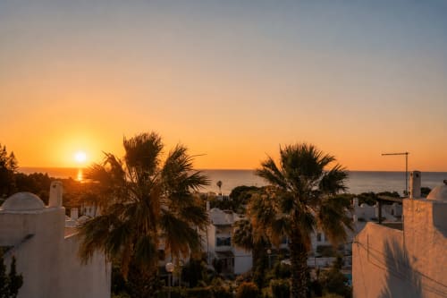 Pôr do sol sobre o mar no apartamento AlgarStays Alporchinhos com palmeiras, edifícios brancos e terraço com vista para o oceano