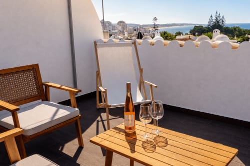 Terraço ensolarado com espreguiçadeiras de madeira, garrafa de vinho e copos na mesa, paredes brancas de privacidade, vista da cidade do Algarve