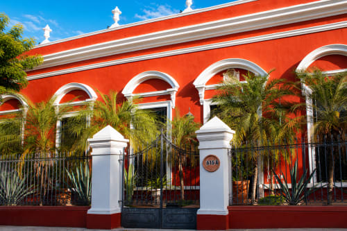 Entrada del hotel boutique Villa Merida con fachada colonial roja vibrante, arcos blancos y paisajismo tropical con palmeras en Mérida