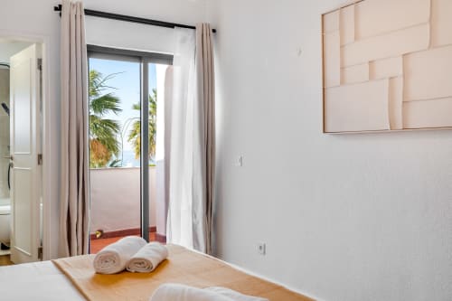 Quarto iluminado com terraço com vista para o mar no apartamento AlgarStays Algarve, toalhas enroladas na cama, palmeiras visíveis através do vidro