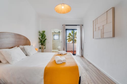 Quarto luminoso com cabeceira de madeira, roupa de cama branca, manta laranja, portas com vista para o mar, arte moderna e iluminação aconchegante com luminárias pendentes