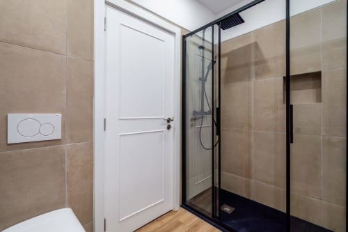 Banheiro moderno com azulejos bege, chuveiro de vidro, porta branca e vaso sanitário no apartamento AlgarStays Algarve.