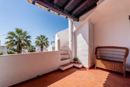 Terraço ensolarado do apartamento AlgarStays Algarve com vista para o mar, cadeira de vime, paredes rosa e palmeiras