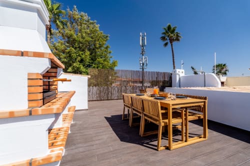 Terraço ensolarado com mesa de jantar em madeira, paredes brancas, palmeiras e vista para o mar em AlgarStays Algarve