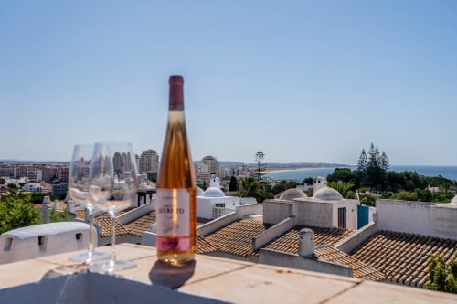 Taça de vinho rosé na varanda do apartamento AlgarStays no Algarve com vista para o mar, edifícios com telhados brancos e praia costeira