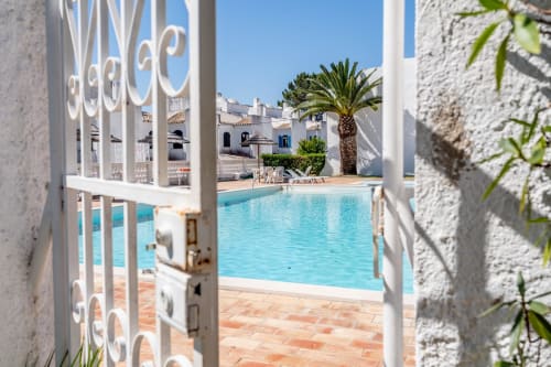 Villa mediterrânea com piscina vista através de portão branco, palmeira, pátio em terracota, edifícios caiados, Algarve