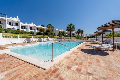 Piscina ensolarada no apartamento AlgarStays Algarve com edifícios brancos, palmeiras, espreguiçadeiras e vista para o mar